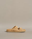 The Suede Hutton Slide Sandal, a tan suede slide with two buckle straps and a ridged sole, is shown against a plain beige background.