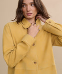 A woman with wavy brown hair wears the Suede Lennox Jacket in a honey shade and gold rings, looking to the side with one hand touching her neck against a beige background.