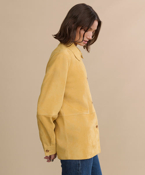 A person with shoulder-length brown hair, dressed in the Suede Lennox Jacket and blue jeans, stands sideways against a plain beige background, looking downward.