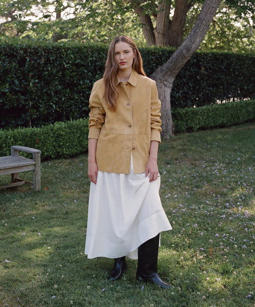 A woman stands on grass in a garden, wearing the Suede Lennox Jacket over a long white dress and black boots. A wooden bench, green hedges, trees, and scattered petals are visible in the background.