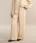 A person wearing a loose, beige, long-sleeve button-up shirt and the Summer Demi Pant stands with one hand in their pocket. Only the lower part of their face is visible, and they have on black woven shoes.