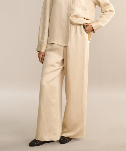 A person wearing a loose, beige, long-sleeve button-up shirt and the Summer Demi Pant stands with one hand in their pocket. Only the lower part of their face is visible, and they have on black woven shoes.