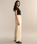 A woman with long dark hair stands sideways against a plain light beige background, wearing a black short-sleeve top, black sandals, and the Summer Demi Pant—loose linen pants featuring an elastic waistband.