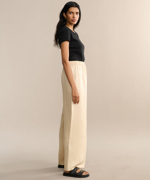 A woman with long dark hair stands sideways against a plain light beige background, wearing a black short-sleeve top, black sandals, and the Summer Demi Pant—loose linen pants featuring an elastic waistband.