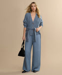A woman with wavy blonde hair poses against a beige background, dressed in a light blue cardigan and matching Summer Demi Pant. She accessorizes with a black handbag and black shoes.