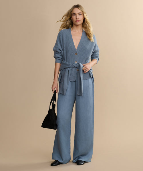 A woman with wavy blonde hair poses against a beige background, dressed in a light blue cardigan and matching Summer Demi Pant. She accessorizes with a black handbag and black shoes.