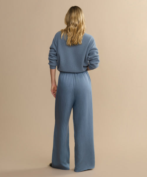 A person with long blonde hair stands facing away, wearing a blue ribbed sweater and the Summer Demi Pant, which features a loose fit and linen texture, set against a plain beige background.
