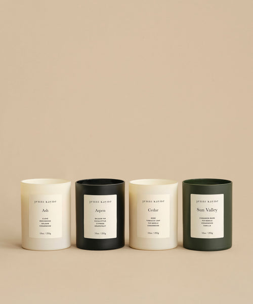 Four Sun Valley Glass Candles—two white, one dark green, one light green—sit in a row on a beige surface. Each minimalist glass jar displays a simple white label with the scent name and branding.