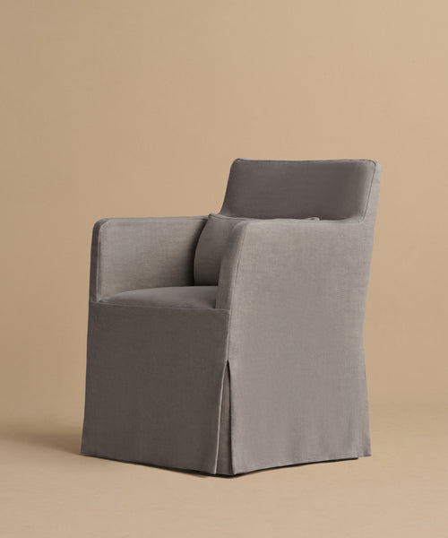The Sunset Dining Arm Chair features a modern design with a grey slipcover, square arms, and a slightly reclined backrest, shown against a plain beige background.