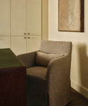 A beige upholstered armchair with a matching lumbar pillow, positioned in a warm-toned room next to a dark wood desk and in front of cream-colored cabinets.