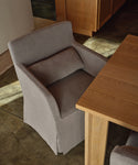 A light gray upholstered dining armchair with a matching back cushion, shown beside a wooden table in a real interior setting with concrete floor and wood cabinetry.