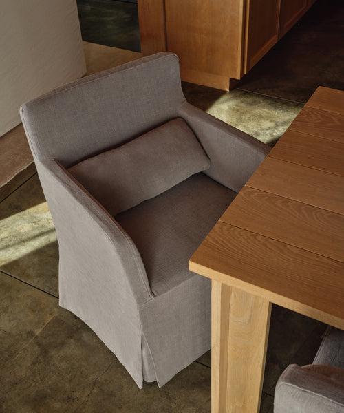 A light gray upholstered dining armchair with a matching back cushion, shown beside a wooden table in a real interior setting with concrete floor and wood cabinetry.