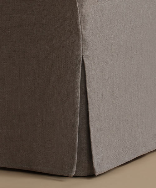 Close-up of a slipcovered upholstered chair or ottoman corner, showing a dark taupe side panel and a light taupe seat/back area with a vertical slit seam, on a beige floor.