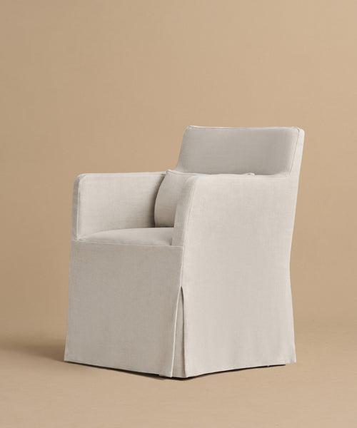 The Sunset Dining Arm Chair features a light beige linen upholstery, high back, straight arms, and a tailored skirt that covers the legs, shown against a plain tan background.