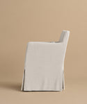 A side view of the Sunset Dining Arm Chair features light gray linen upholstery, a straight back, and a long, tailored skirt covering the legs, set against a plain beige background.