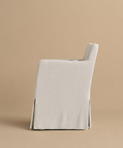 A side view of the Sunset Dining Arm Chair features light gray linen upholstery, a straight back, and a long, tailored skirt covering the legs, set against a plain beige background.