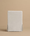 The Sunset Dining Arm Chair is shown from behind, featuring a beige linen tailored slipcover with neat seams and side vents, and a straight back, set against a light tan background.