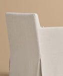 The Sunset Dining Arm Chair, featuring a beige linen slipcover and straight backrest, is set against a plain light brown background.