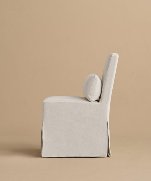 The Sunset Dining Chair features a modern armless design with light beige linen upholstery, a square seat, tall back, and round lumbar cushion, set against a tan background that showcases classic California style.