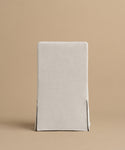 A simple, straight-backed Sunset Dining Chair with a light beige linen slipcover inspired by California design, featuring two front slits near the bottom corners, shown against a plain tan background.