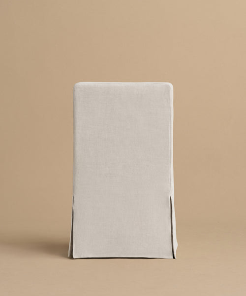 A simple, straight-backed Sunset Dining Chair with a light beige linen slipcover inspired by California design, featuring two front slits near the bottom corners, shown against a plain tan background.