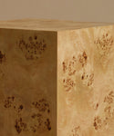A close up of a square burl wood side table standing upright on a beige surface against a solid beige background.