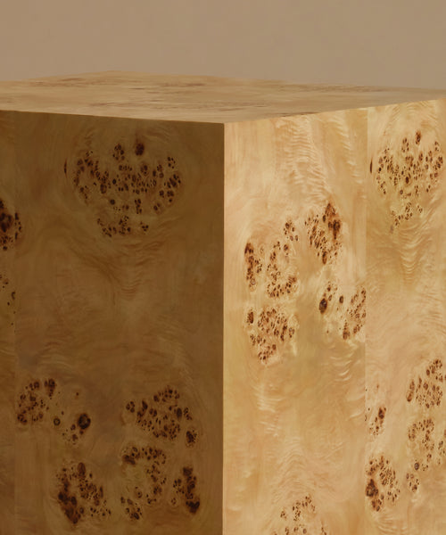 A close up of a square burl wood side table standing upright on a beige surface against a solid beige background.