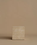 A square travertine side table, standing upright on a matching beige surface against a solid beige background.