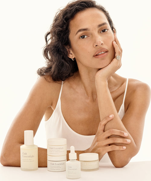 A woman with wavy dark hair and a neutral expression rests her face on her hand. She wears a white tank top and sits behind The Routine by Oak Essentials, a 5-step glowing skin secret, arranged on a white surface.
