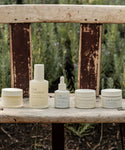 A row of Oak Essentials The Routine skincare products, a nourishing 5-step set, sits on a weathered wooden bench outdoors amid blurred greenery and lavender plants.