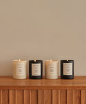 Four lit Topanga Glass Candles in black and cream jars line a wooden surface against a beige wall. Each hand-poured Los Angeles candle has a minimalist label, reflecting natural scents and elegant design.