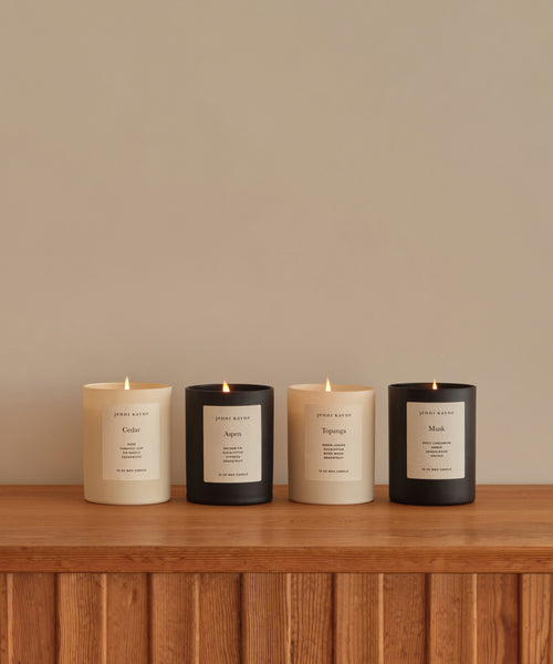 Four lit Topanga Glass Candles in black and cream jars line a wooden surface against a beige wall. Each hand-poured Los Angeles candle has a minimalist label, reflecting natural scents and elegant design.