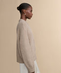A woman with hair in a low bun and pearl earrings wears the Tove Crewneck, a beige cable knit sweater, paired with a light skirt, standing in profile against a plain beige background.