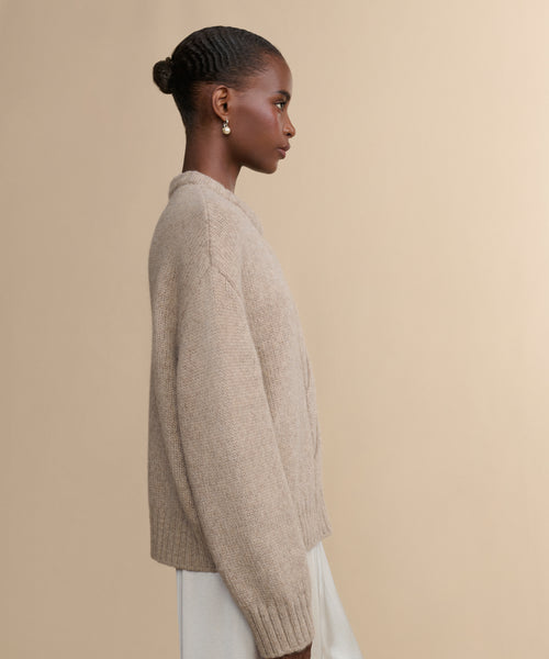 A woman with hair in a low bun and pearl earrings wears the Tove Crewneck, a beige cable knit sweater, paired with a light skirt, standing in profile against a plain beige background.