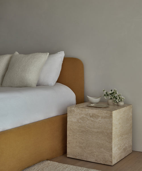 A minimalist bedroom featuring the Sur Side Table in solid travertine, a mustard upholstered bed with white bedding, and small ceramic vessels with a sprig of flowers against a light gray wall.