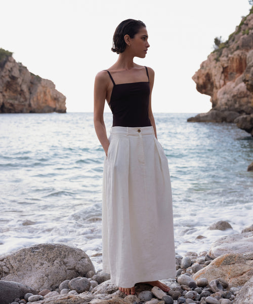 A woman stands barefoot on a rocky beach, wearing the Tre One Piece with waves and cliffs behind her under a bright sky.