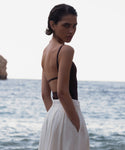 A person with short hair stands by the sea in a dark Tre One Piece, hands in pockets, glancing over their shoulder at the camera. The bright background shows sparkling water and a rocky formation.