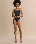 A barefoot woman with curly hair stands against a beige background, wearing the Tre One Piece swimsuit. She faces forward with a neutral expression, her arms relaxed at her sides.