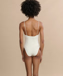 A person with short curly hair is seen from behind, wearing the Trentasei One Piece swimsuit with thin straps against a plain beige background.