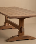 The Trestle Dining Table features a rectangular plank-style top, sturdy trestle legs, and a natural wood finish, shown against a plain beige background.