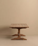 The Trestle Dining Table features a handcrafted rectangular top and sturdy central pedestal base, with visible wood grain and clean, simple lines for a timeless look, set against a plain beige background.