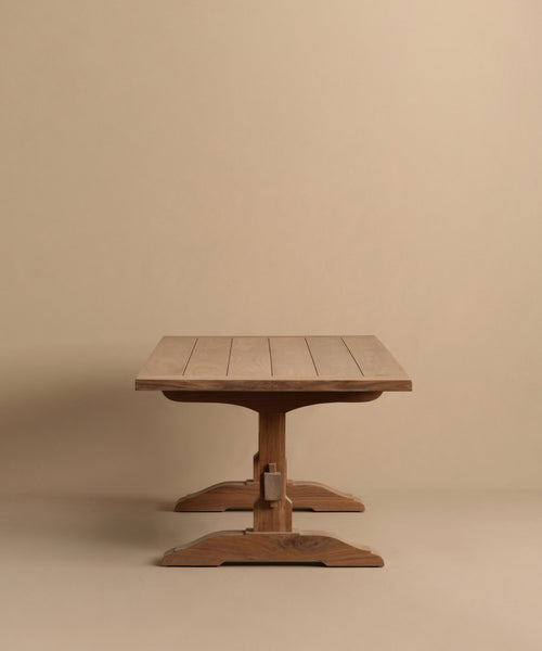 The Trestle Dining Table features a handcrafted rectangular top and sturdy central pedestal base, with visible wood grain and clean, simple lines for a timeless look, set against a plain beige background.