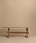 The Trestle Dining Table features a handcrafted rectangular top and sturdy crossbeam supports, displayed against a plain beige background.