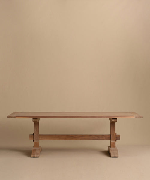 The Trestle Dining Table features a handcrafted rectangular top and sturdy crossbeam supports, displayed against a plain beige background.