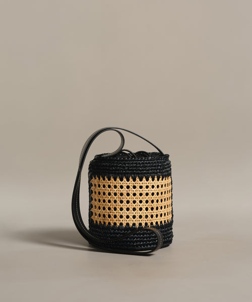 The Tropez Bucket Bag is a small cylindrical handbag with a woven black base and patterned beige rattan middle, featuring a long black strap and handcrafted cane design. Shown against a plain light gray background.