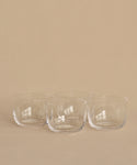 Four clear, round hand-blown bowls from the Universal Low Glass Set of 4 sit in two rows on a beige surface and background. The empty potassium glass bowls feature smooth, minimalist designs inspired by Vincent Van Duysen.