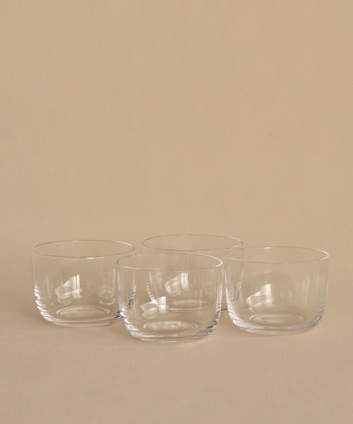 Four clear, round hand-blown bowls from the Universal Low Glass Set of 4 sit in two rows on a beige surface and background. The empty potassium glass bowls feature smooth, minimalist designs inspired by Vincent Van Duysen.
