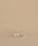 A clear, hand-blown tumbler from the Universal Low Glass Set of 4 rests on a beige surface with a matching background, highlighting Vincent Van Duysen’s minimalist style.