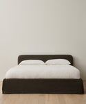 The Cove Bed Slipcover, featuring a dark upholstered frame and headboard, is dressed in white Jet Linen bedding with two white pillows. It sits on a light wood floor against a plain, light-colored wall.