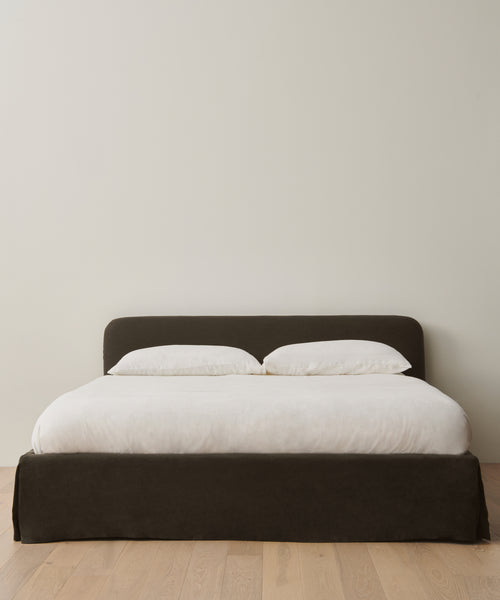 The Cove Bed Slipcover, featuring a dark upholstered frame and headboard, is dressed in white Jet Linen bedding with two white pillows. It sits on a light wood floor against a plain, light-colored wall.